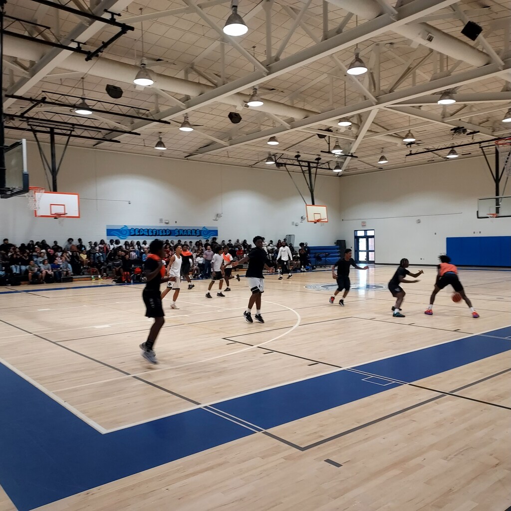 An indoor basketball court with blue markings. A group of people are playing basketball with others watching.