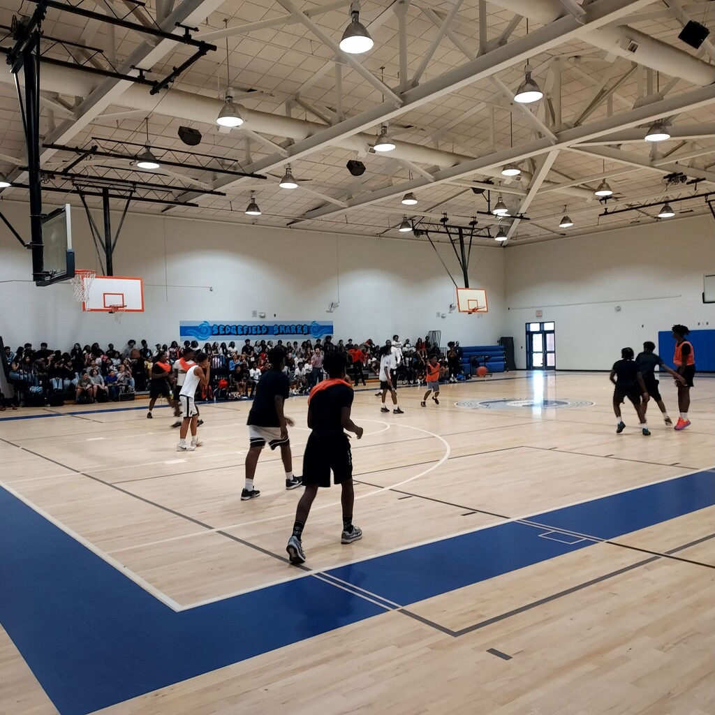 Indoor basketball court with players in action, wooden floor, and spectators seated on the side.