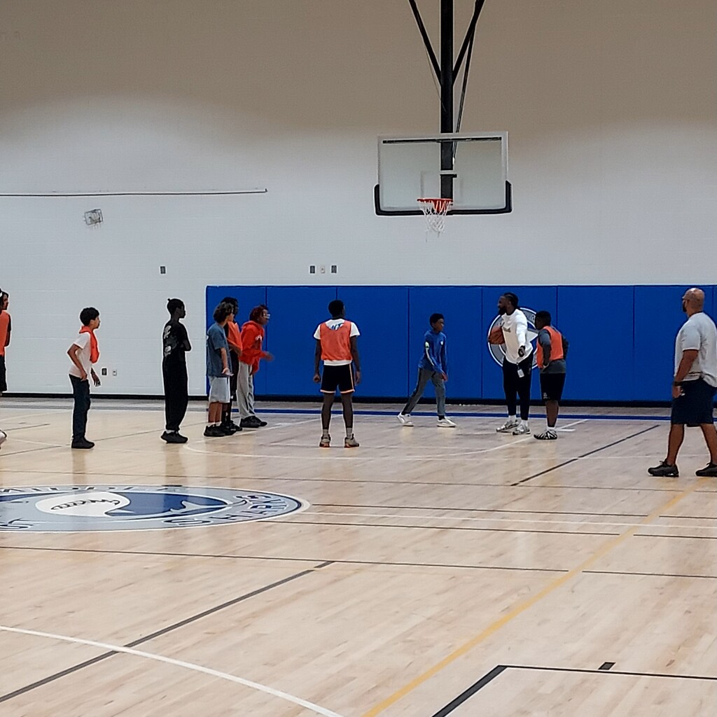 Indoor basketball court with a group of people. Some wear sneakers, others wear jerseys. A basketball hoop on the wall.