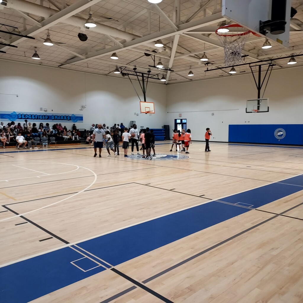 An indoor basketball court with spectators on the side, players standing in groups, and two basketball hoops.