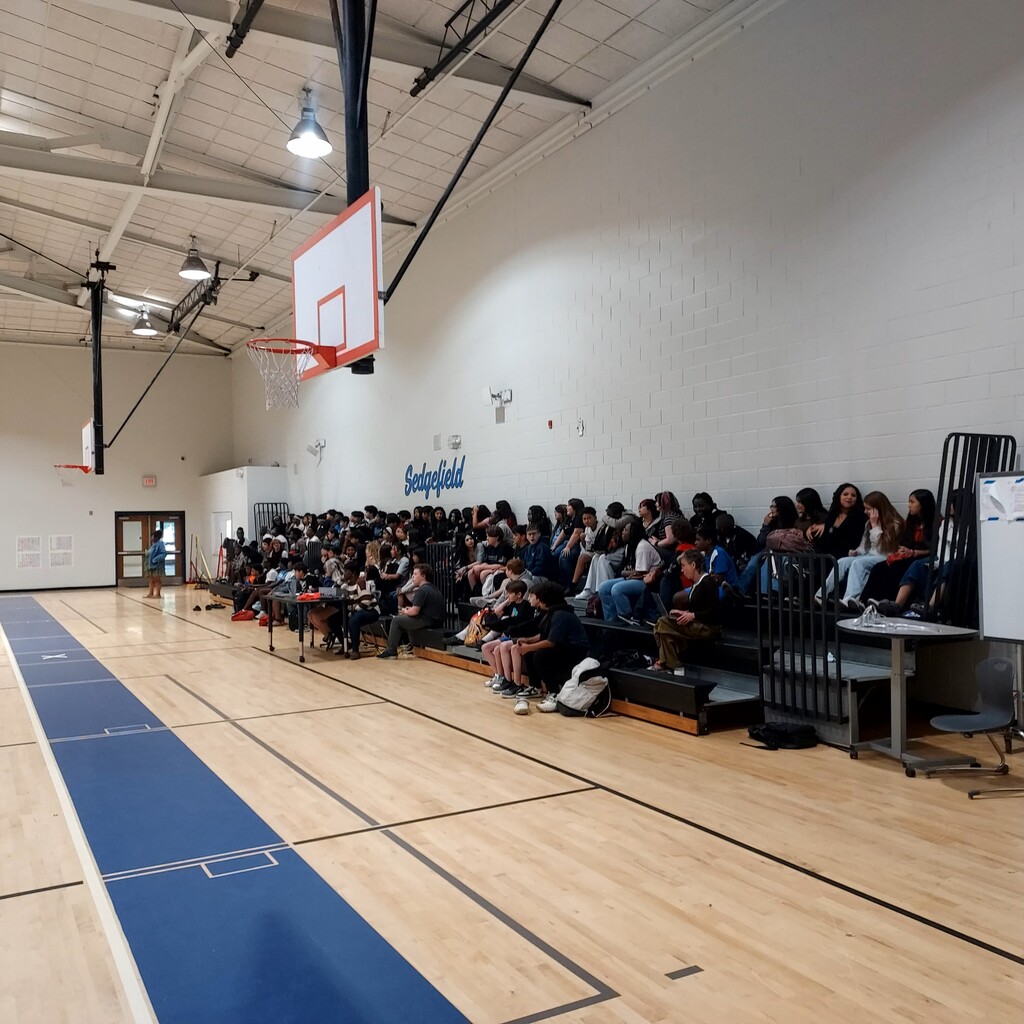 Interior of an indoor basketball court. A crowd sits on benches, watching an event. A basketball hoop is mounted on a wall.