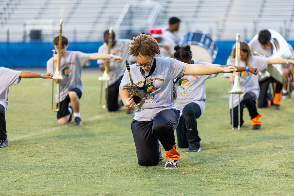 A marching band practices on a field. Members are holding musical instruments. Some wear gray shirts, black pants, and orange socks.