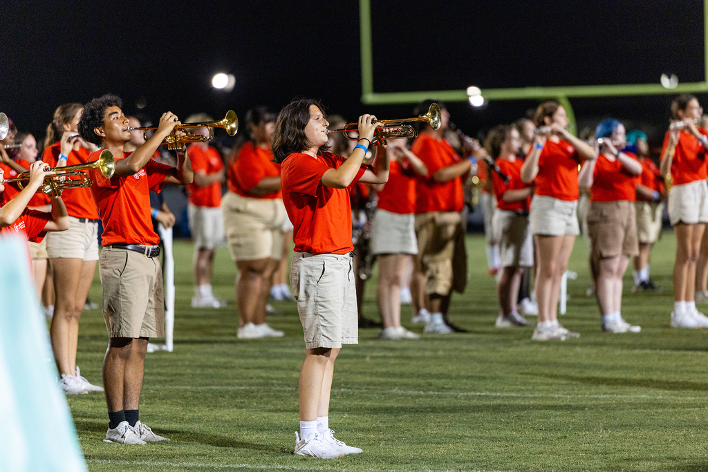 A group of musicians wearing orange shirts and khaki shorts play brass instruments on a football field at night.