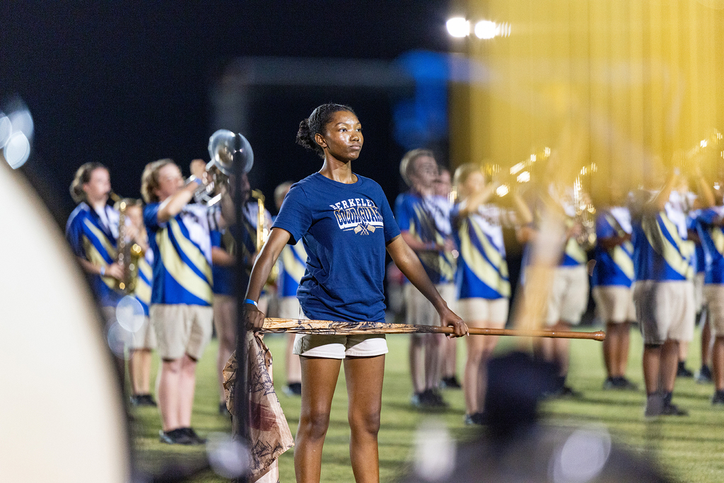 Woman with baton stands on field, holding flag. Other band members play instruments in background.