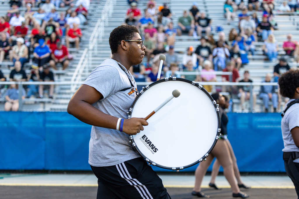 A man plays a drum in a marching band, with others in the background and an audience seated in bleachers.