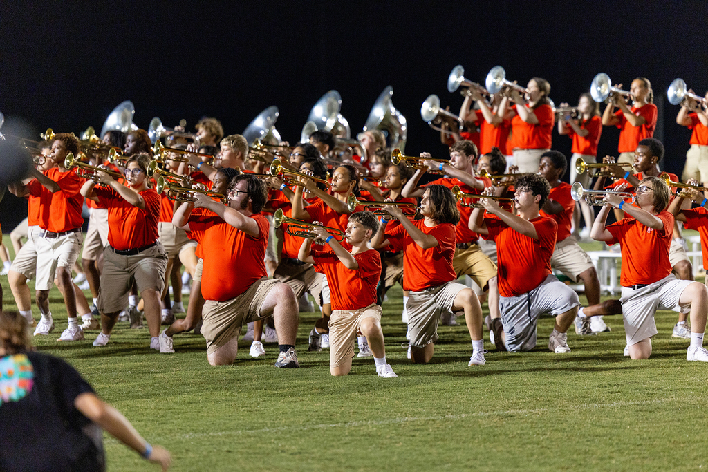 Musicians wearing red shirts and khaki pants play instruments while kneeling on a grass field.