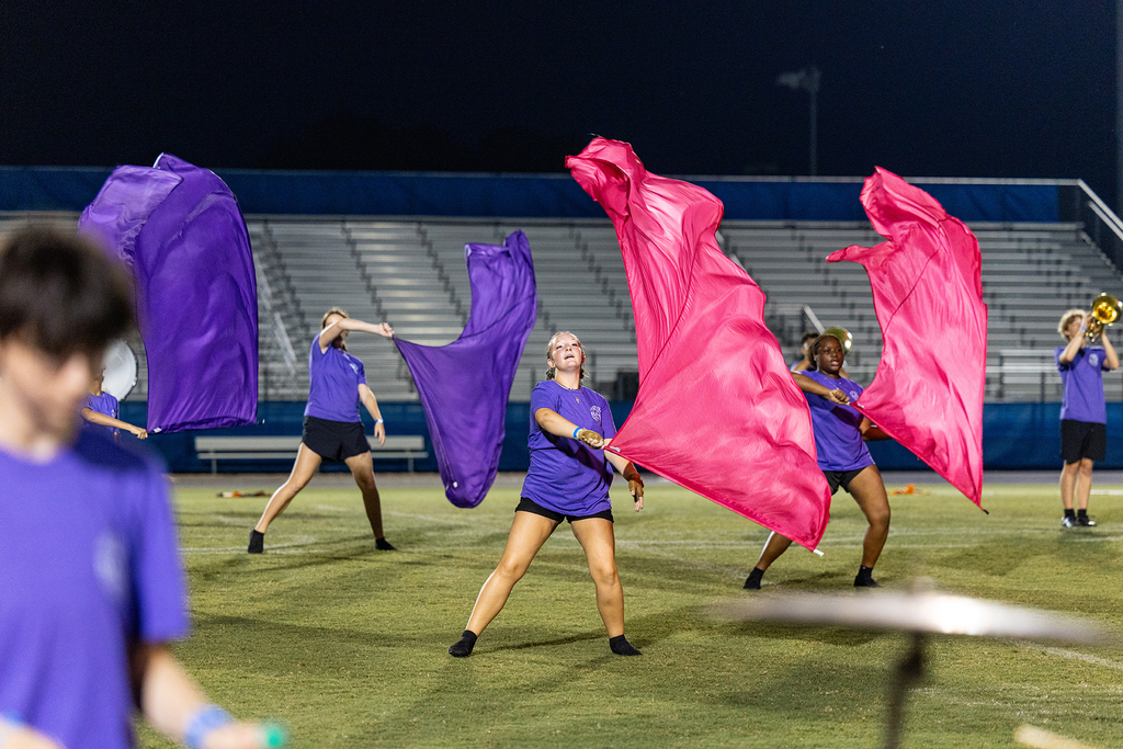 Five people in matching purple and pink outfits perform on a field at night. They are waving fabric.