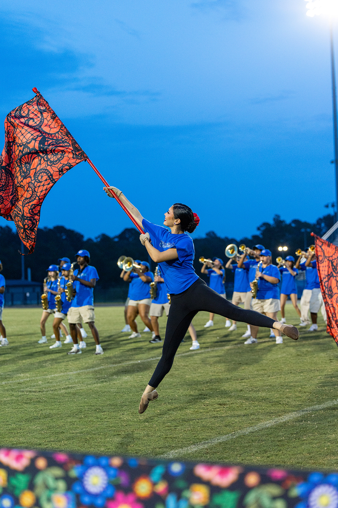 A woman holds a flag as she jumps on a football field with a marching band in the background.