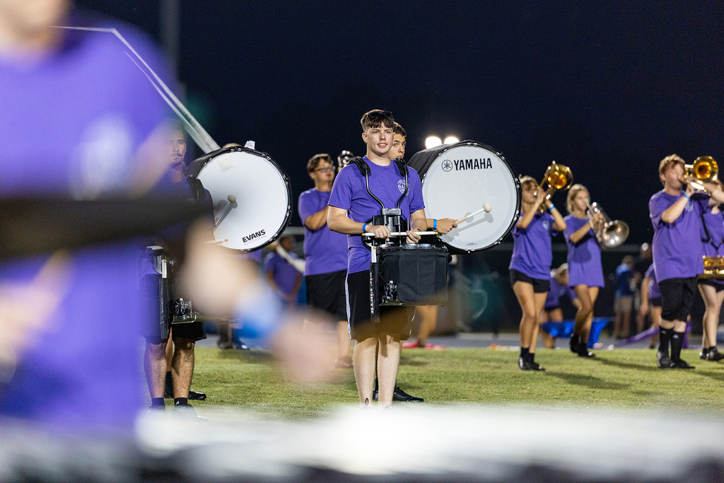 A group of people in purple uniforms play instruments on a field at night. Some are on the ground and others are standing.