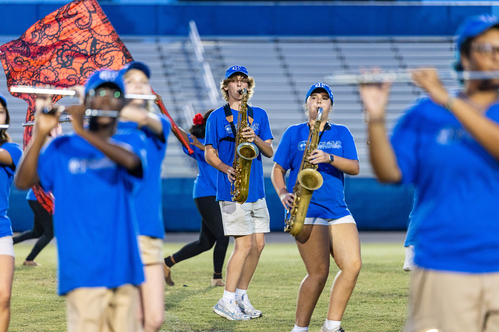 A school band practices outdoors. Several students play saxophones, flutes, and a flag. They wear matching blue shirts.