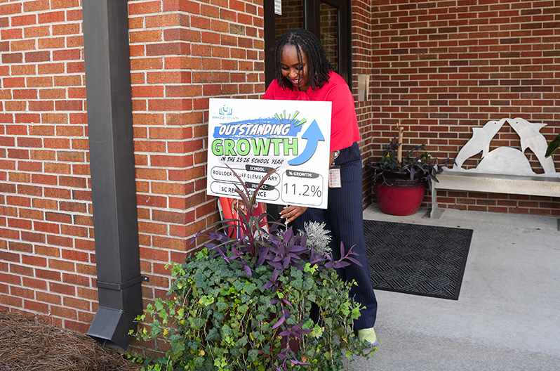 Person holds sign with text "Outstanding Growth" in front of brick building with greenery and a bench.