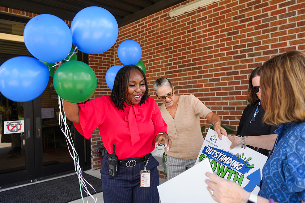 A woman holding balloons stands next to another holding a sign reading "Outstanding Growth" in front of a brick wall.