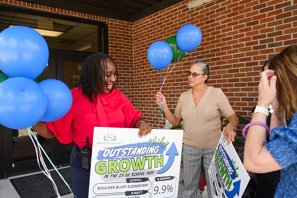 Four women, one holding a sign reading "Outstanding Growth," stand outside a brick building. Balloons are held by three women.