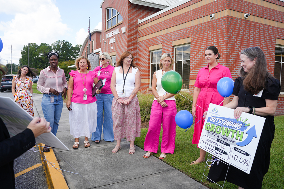 Nine women in pink stand on a sidewalk in front of a building. One holds a sign.