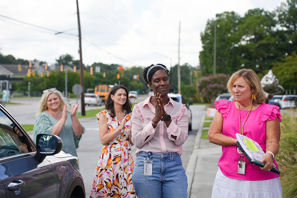 Five women on a road. One woman clapping hands and the other holding a folder. Vehicles and trees in the background.