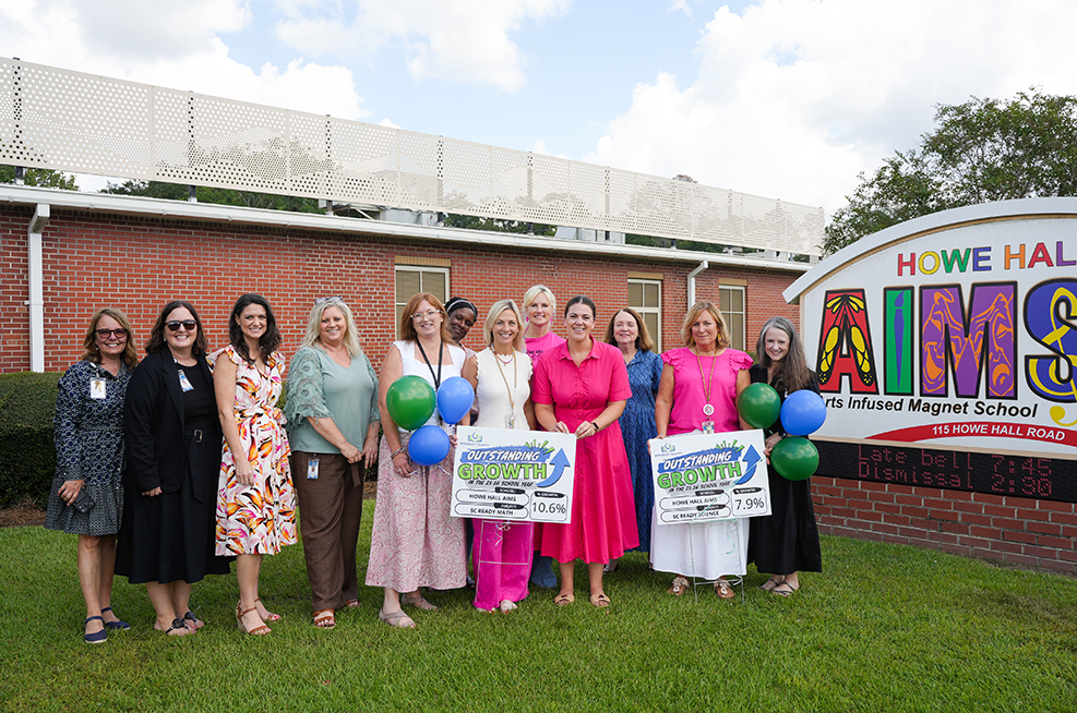 Eleven people stand on grass in front of a sign reading "AIMS Howe Hall". They hold green balloons.