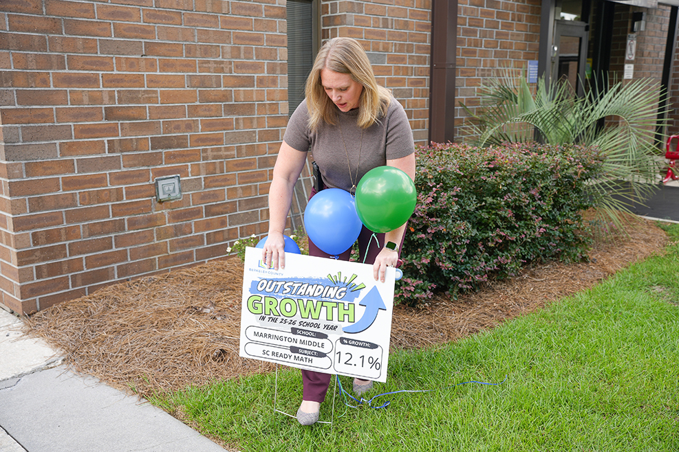 A person holds a sign with balloons and a graph, standing on grass outside a brick building.