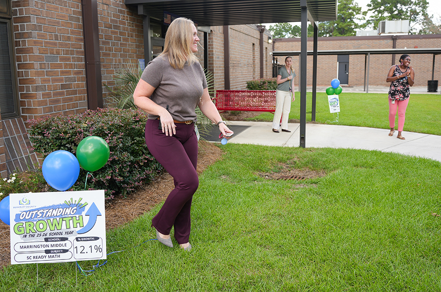 Woman in maroon pants and gray top near a sign on the lawn. Two other people walk in the background.