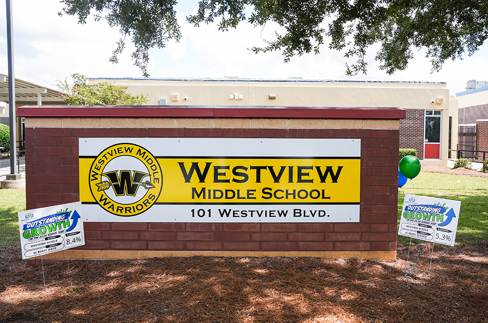 Entrance to Westview Middle School with a yellow and white sign. Three signs display on the ground. A tree casts a shadow over the sign.