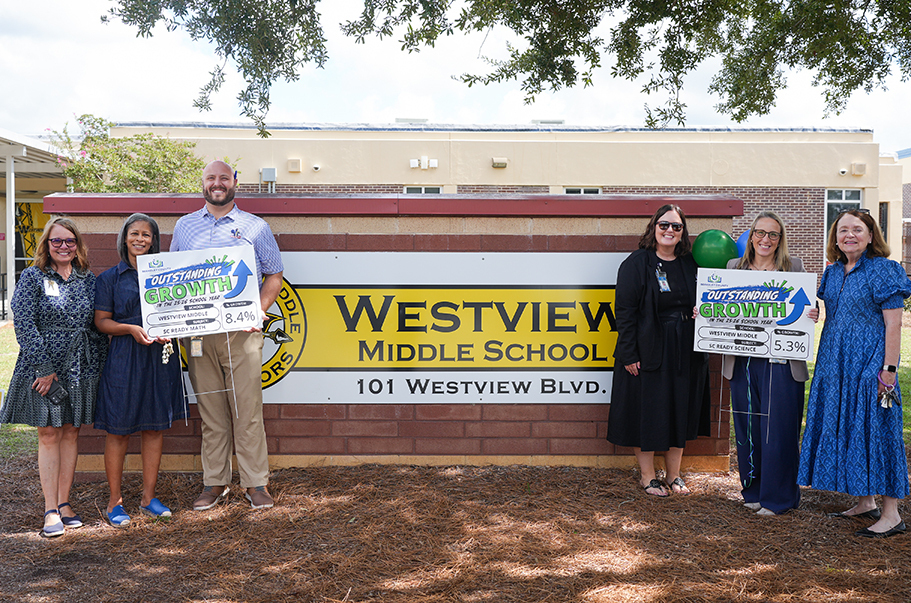 Five people stand before a brick wall, holding signs. The sign reads "Westview Middle School." A building is in the background.