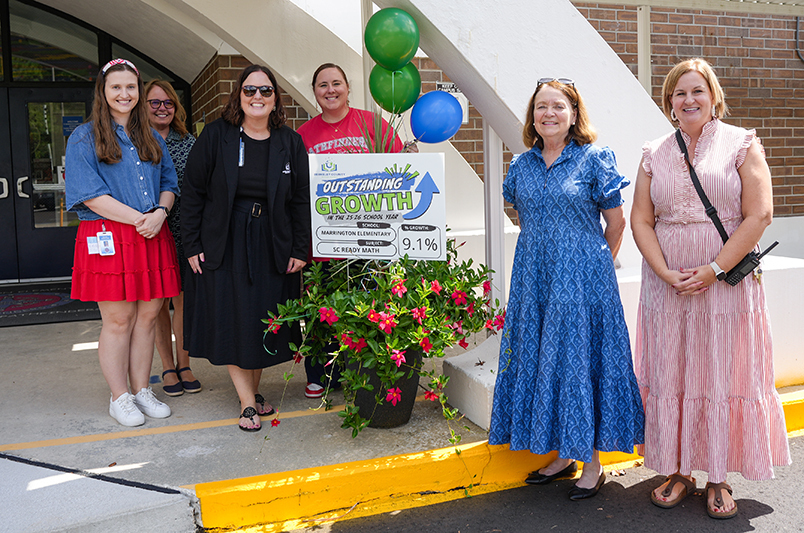 Group of six adults posing near a flowerpot and sign. One holds a sign that reads "Community Growth 9.1%".