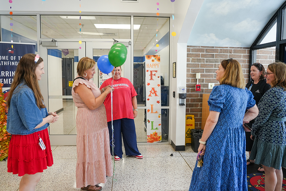 Several people stand in a hallway, some holding balloons. A sign with Fall text is on the wall.