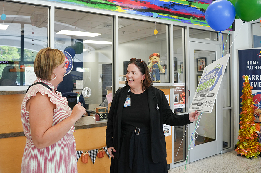 Two women converse at a building's entrance. One holds a sign with colorful graphics. Behind them, balloons, a counter, and framed artwork are visible.