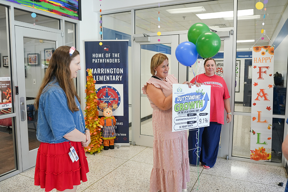 Three women hold balloons and a banner, standing inside a building with glass doors and displays.