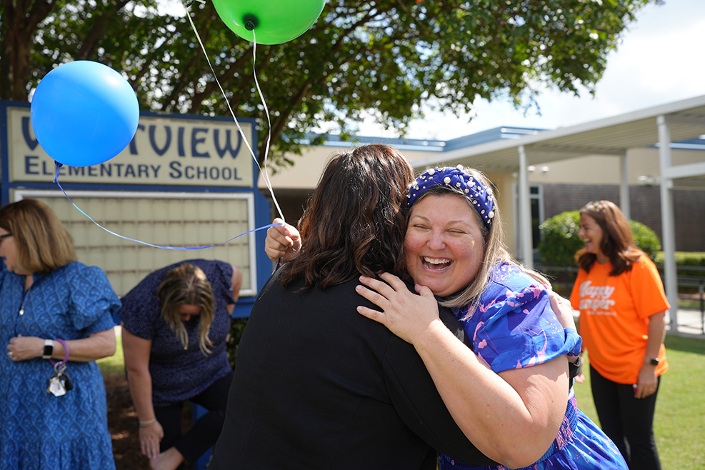 Two women hug in a field, with a third woman holding balloons. Another woman crouches behind a sign.