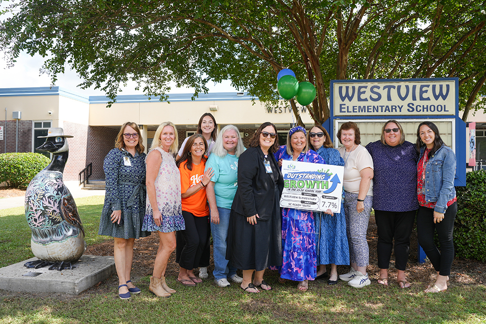 Group of women in summer clothing, standing outdoors in front of a school sign and a decorative goose.