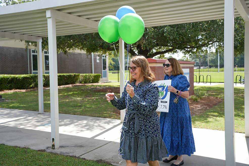 Two women walk, holding balloons, in a covered walkway. One holds a sign. Behind them is a lawn and shrubs.