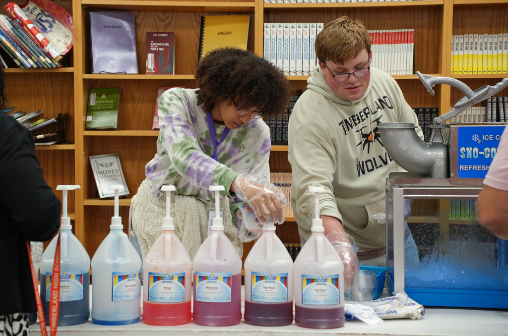 Two students with glasses handle dispenser bottles in a classroom with books and shelves in the background.