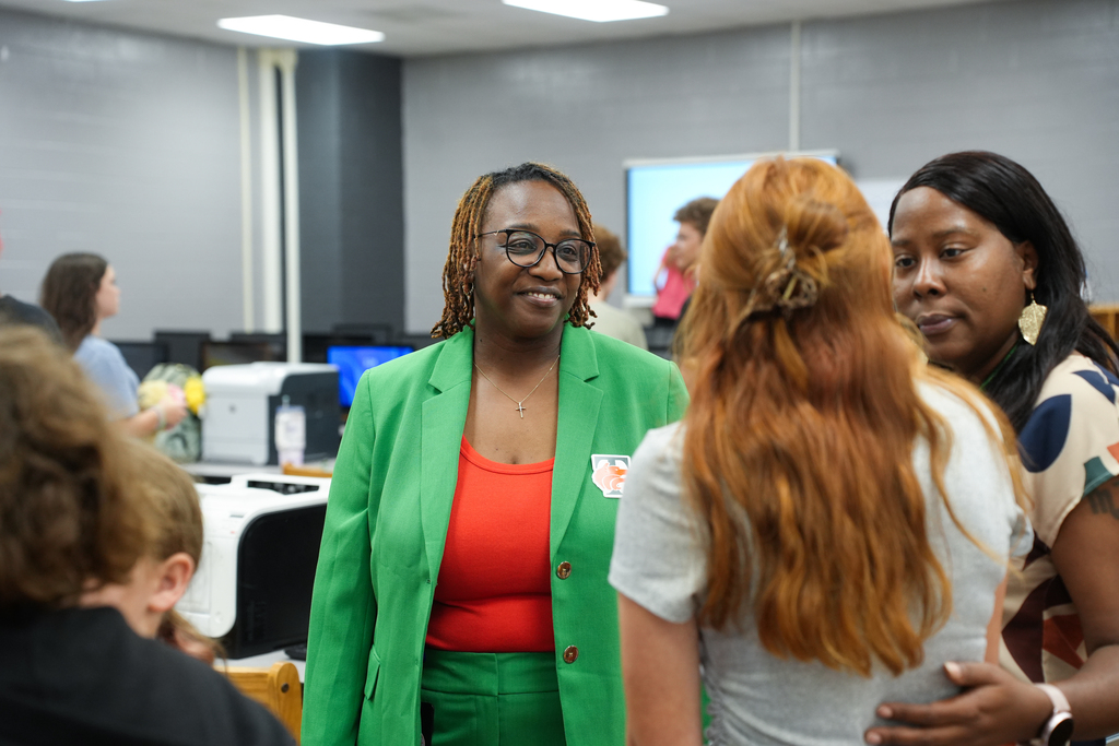 A group of women in an office. One woman wears a green jacket and orange shirt. Another woman has red hair.