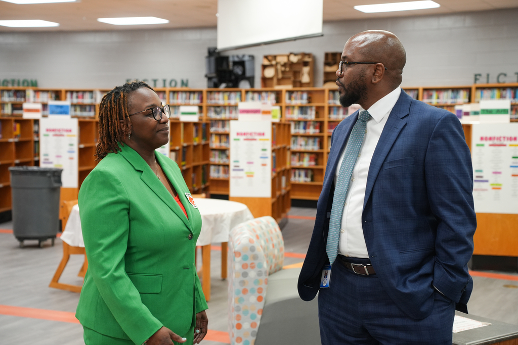 Two individuals in formal attire are engaged in conversation in a library. One wears a green suit, the other, a blue suit.