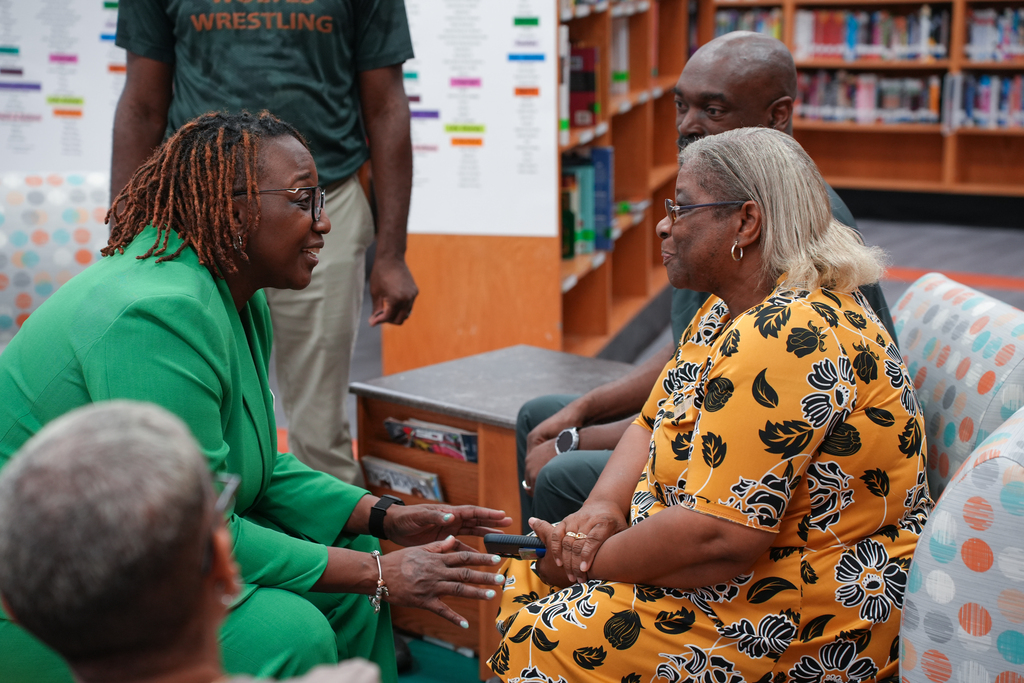Two women sit and talk in a library; one wears green, the other yellow with black patterns. Others sit nearby, and bookshelves and a wrestling poster are in the background.
