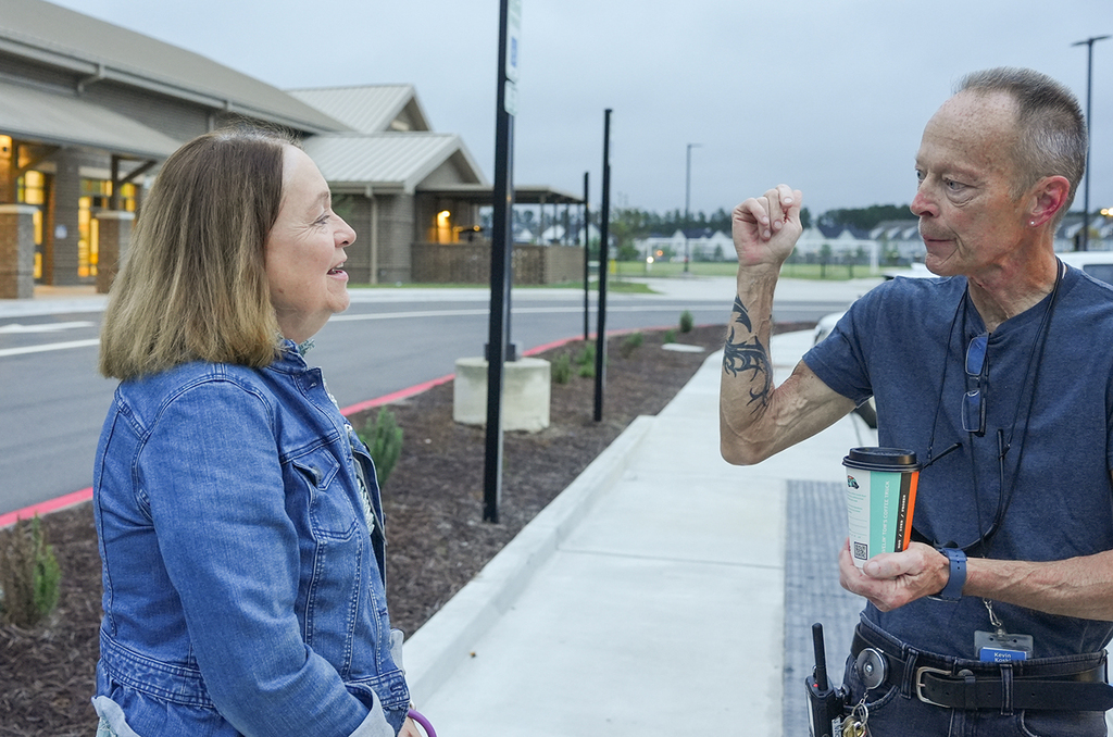 A man and woman in conversation, with the man holding a cup, standing on a sidewalk by a building.