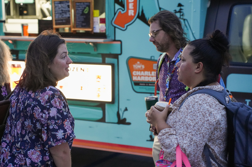 Three people stand by a food truck, one holding a drink, while a menu board displays "Charlie Harbor" on the truck.