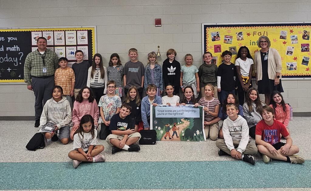 Group of children and two adults in a classroom, posing for a photo. Two adults stand at the front.