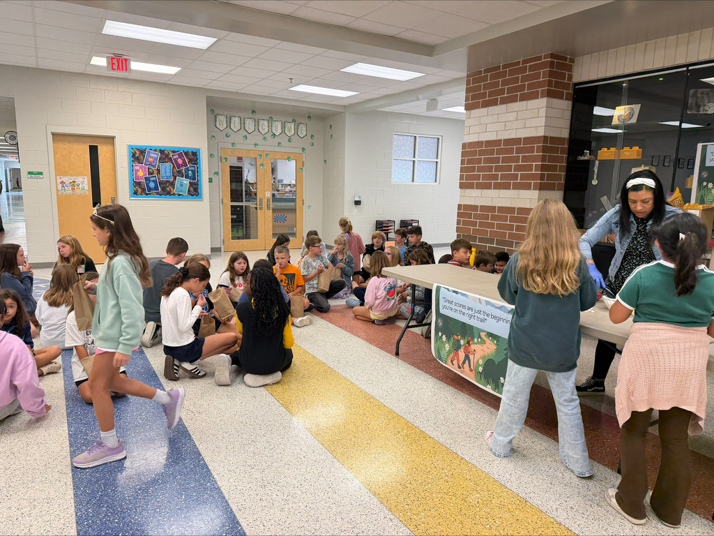 Group of children seated and standing, with two adults in front of a table in a classroom.