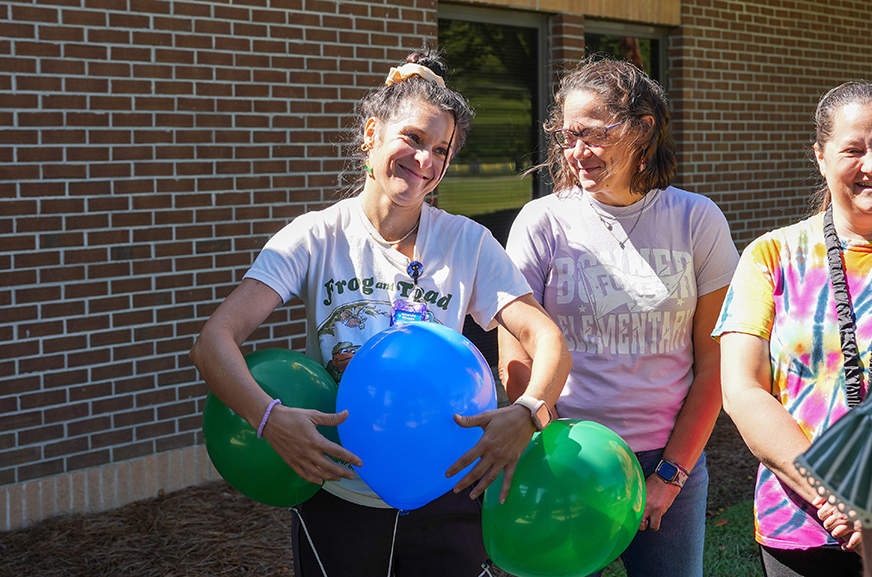 Three women standing, one holding a blue balloon, another with green balloons, near a brick building.