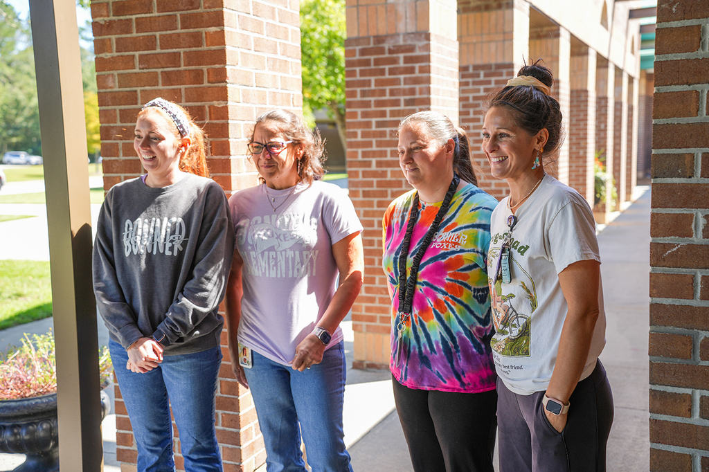 Four women stand in a hallway. They face forward, with a brick wall and an open grassy area behind them.