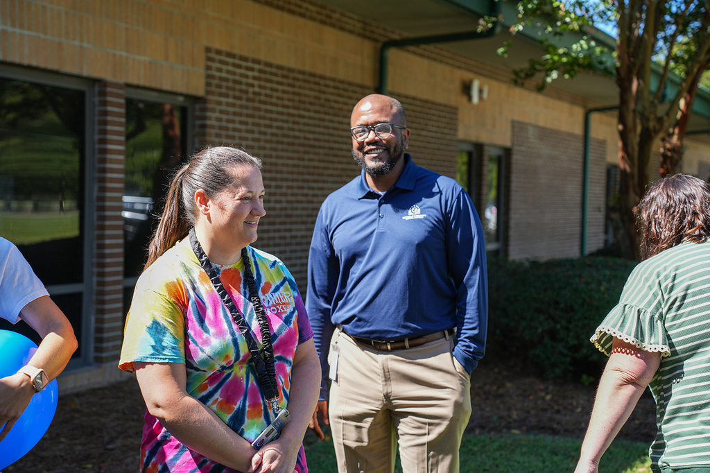 People stand in a group outside a building. A woman in a tie-dye shirt smiles, and a man in a navy polo laughs.