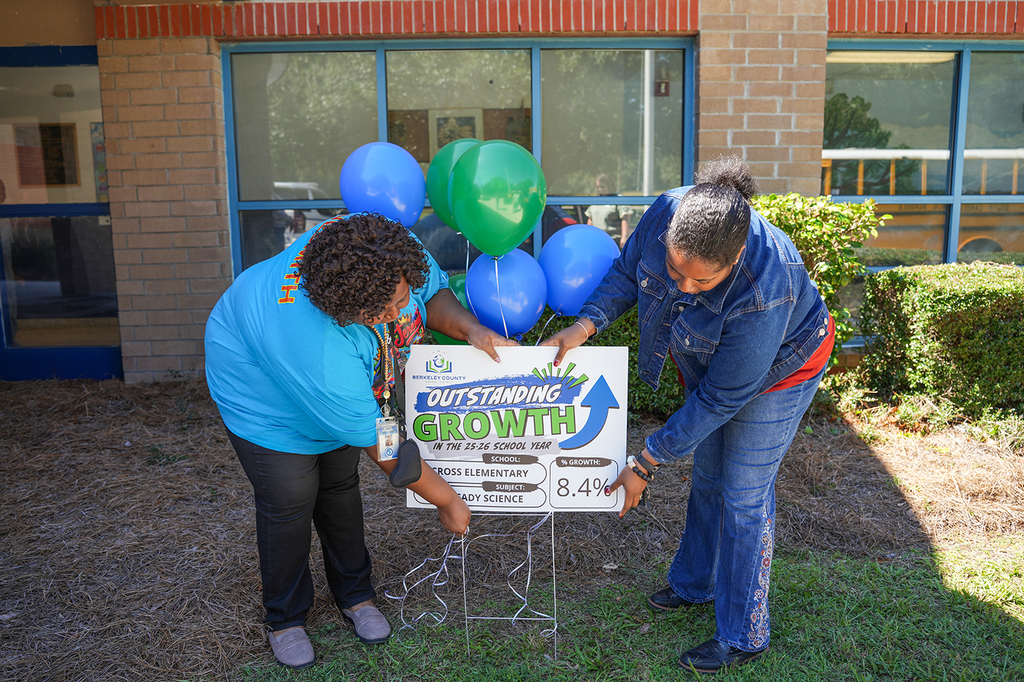Two people hold a board with "Outstanding Growth" text and balloons in front of a building.