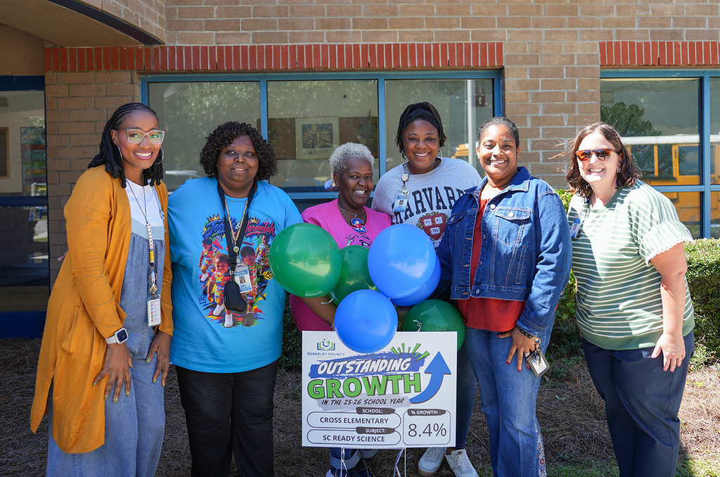 Several people pose with balloons and a sign. Behind them is a school building with a bus parked.