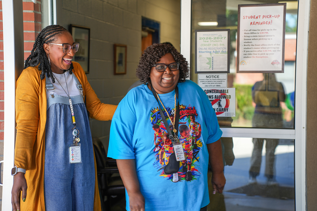 Two women stand outside a building entrance, one smiling. One wears a blue shirt with a colorful design and a lanyard.