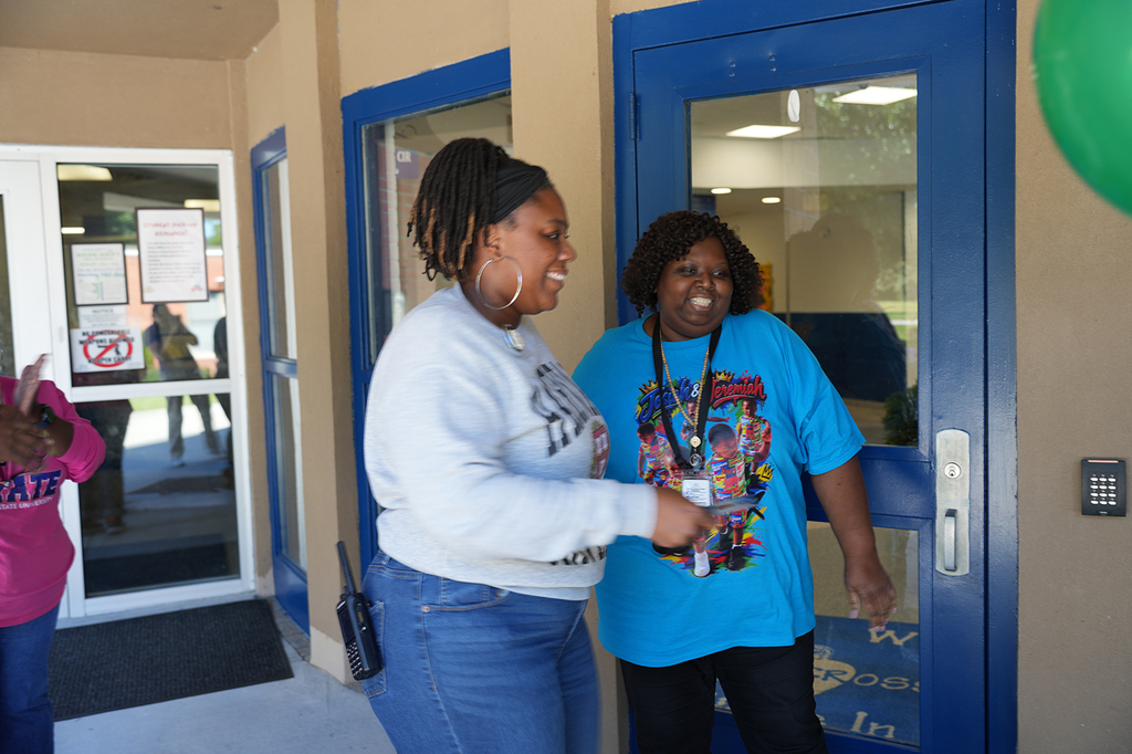 Two women stand at a blue door with a balloon, one wearing white and the other blue, both smiling.