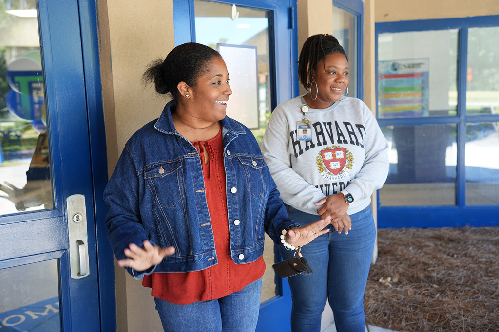 Two women stand before a building entrance; one in denim, the other in a Harvard sweatshirt, both smiling.