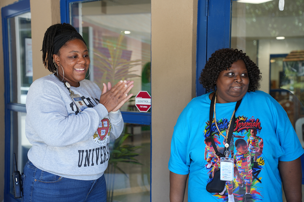 Two women standing together, one clapping hands. One wears a white sweatshirt with "UNIVERSITY" on it; the other has a blue shirt. A stop sign is behind them.
