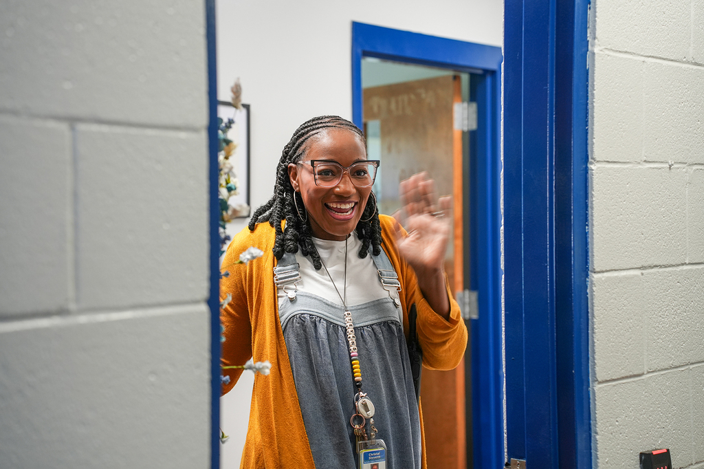 A woman smiles and waves inside a room with blue trim, wearing glasses and a yellow cardigan.