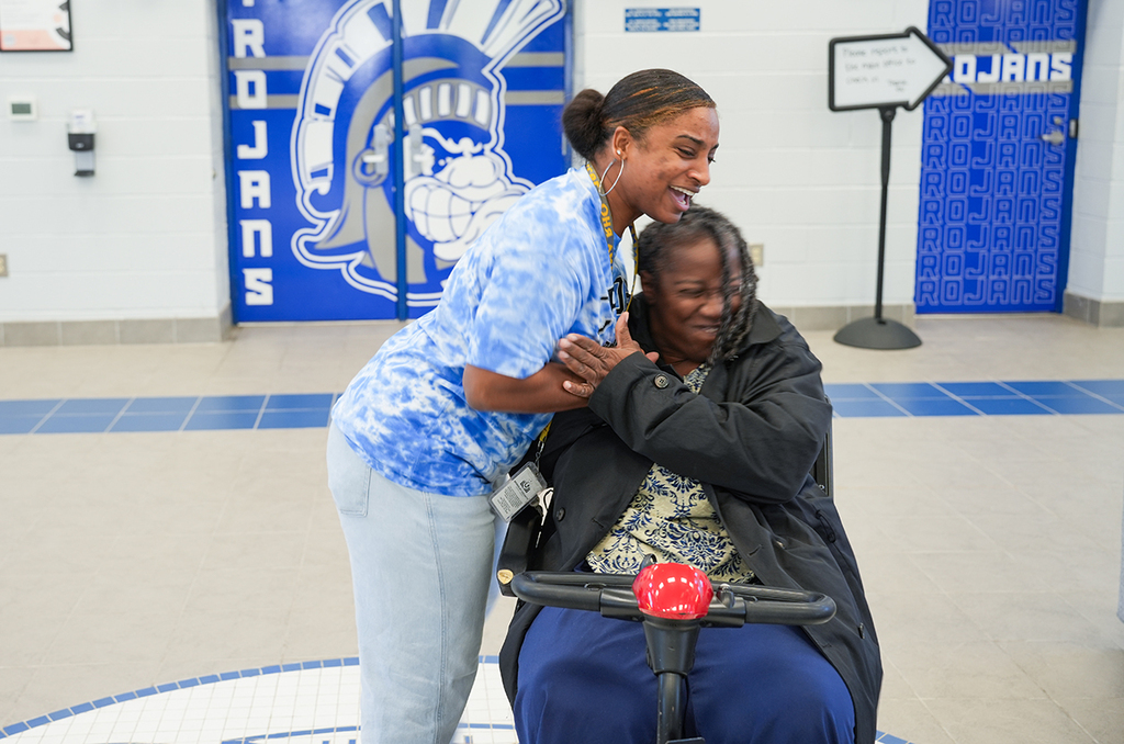 Two women embrace near a school entrance. One in a tie-dye shirt stands, the other sits in a wheelchair.